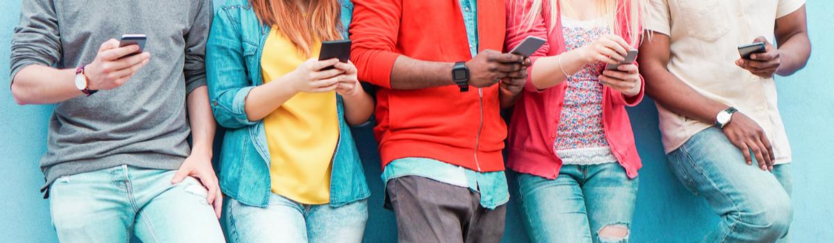 Foto de jovens encostados em uma parede azul. Cada um deles segura um celular nas mãos.